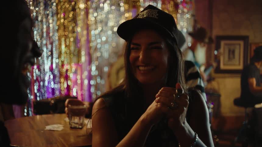 Wearing a baseball cap a woman laughs and uses hand gestures while talking at a table in a bar as a band plays and people dance behind a shimmering tinsel curtain