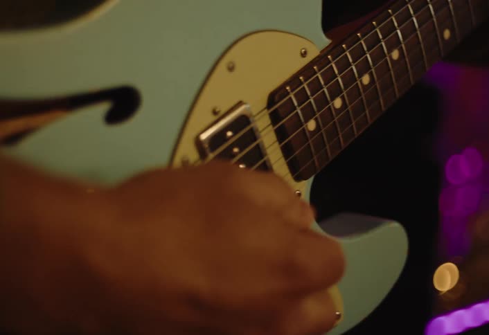 A musicians hands move skillfully over the strings and fretboard of a light blue electric guitar against a dimly lit sparkly background