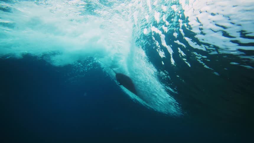Viewed from below the oceans surface a surfer paddles overhead on their board as a wave forms and creates turbulence above the reef