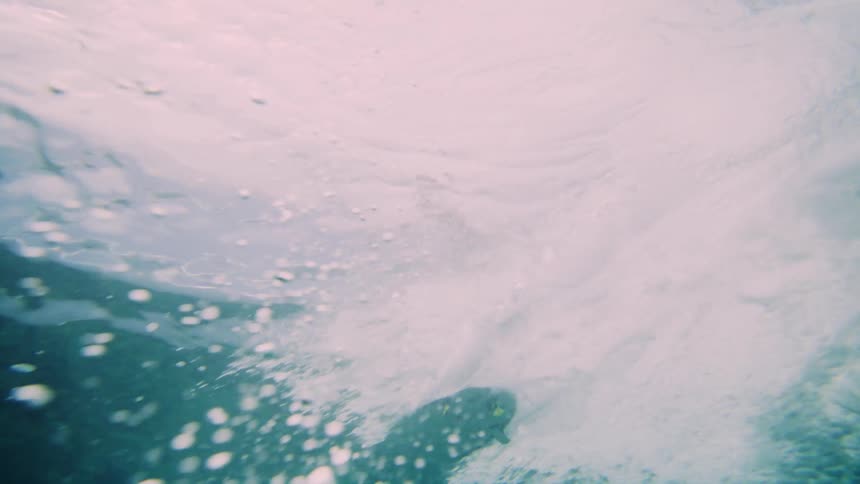 A surfer glides underwater beneath a breaking ocean wave surrounded by churning water and bubbles