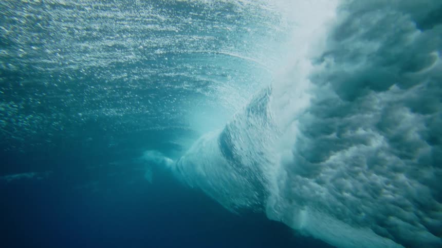 From beneath the surface the turbulent underside of a large ocean wave crashes as a surfboard briefly passes overhead within the churning water and bubbles