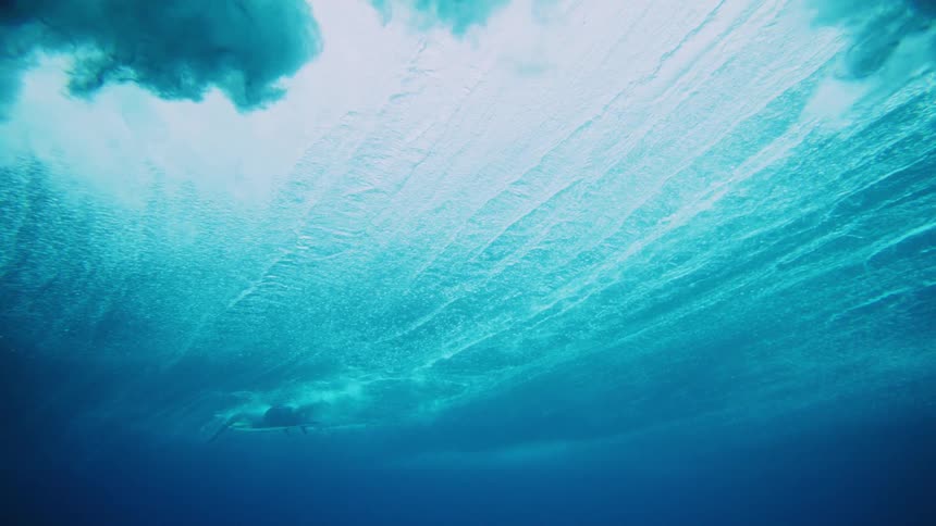 Seen from the ocean floor a scuba diver swims past initially then a large wave passes overhead creating dramatic surface turbulence where a surfer is paddling on their board