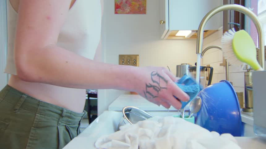 A person wearing a crop top washes a blue mug in a kitchen sink filled with soapy water