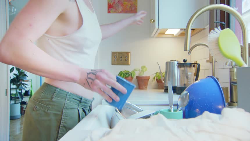 A person uses a white cloth to dry a blue mug next to a sink containing dishes utensils and a dish brush