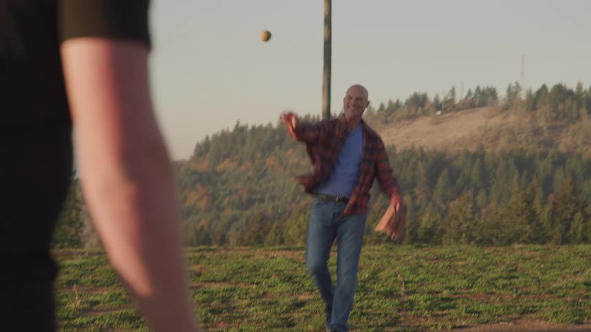 A father smiles as he throws a baseball to his adult son while playing catch outdoors in a grassy field near forested hills