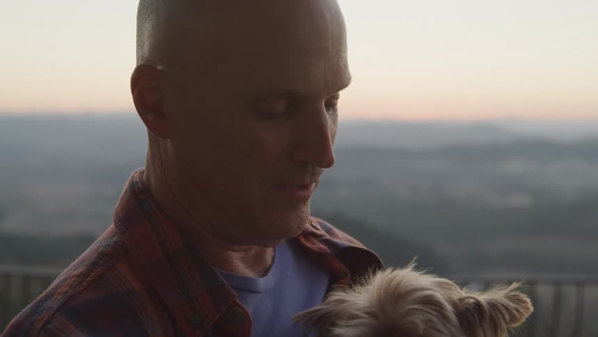 A man holds a small dog outdoors looking down fondly against a hazy sunrise or sunset backdrop