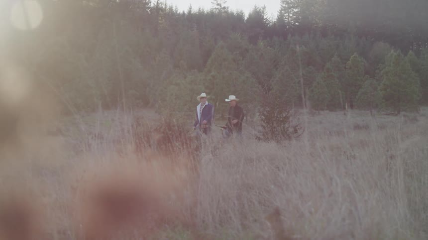 Two men wearing cowboy hats walk through a rural field near a grove of trees on a sunny day