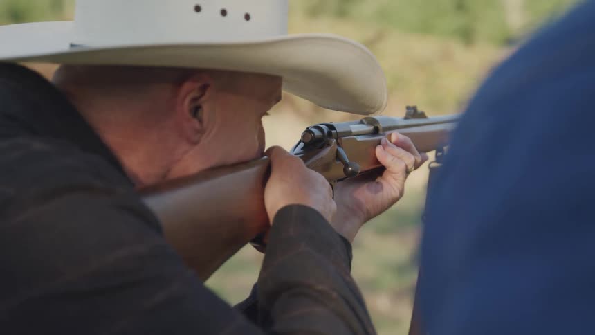 A man in a white cowboy hat takes aim with a boltaction rifle outdoors
