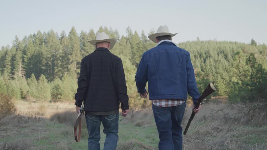 Two hunters are carrying rifles while walking near a forest on a sunny day