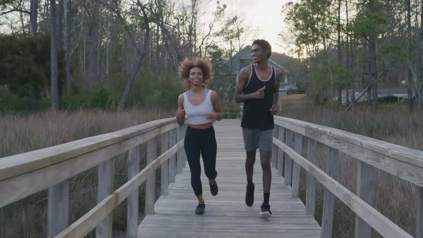 Two people run over a wooden footbridge with a grove of trees and a house in the background