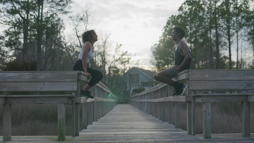 A man and woman in athletic wear sit facing each other on opposite railings of a long wooden boardwalk