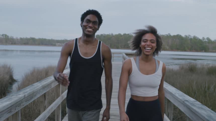 A young couple highfive on a wooden boardwalk surrounded by a marsh with trees and reflective water in the background