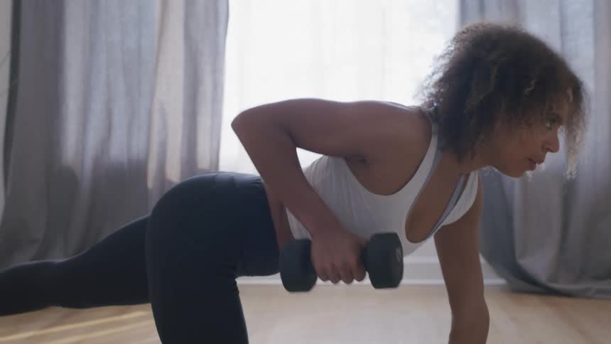 Woman balances on a knee with an outstretched foot and lifts a hand weight while working out at home during the daytime