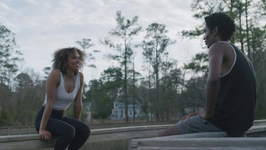 A young man and woman in athletic wear sit opposite each other on a wooden railing outdoors engaged in conversation
