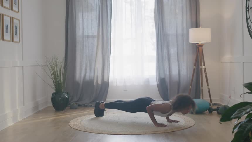 A woman in athletic wear does push ups on a circular rug in a sunlit room with large windows and curtains