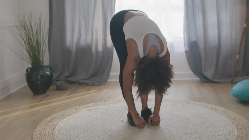 A woman with curly hair performs a standing forward bend stretch on a round rug as part of a home workout in a brightly lit room