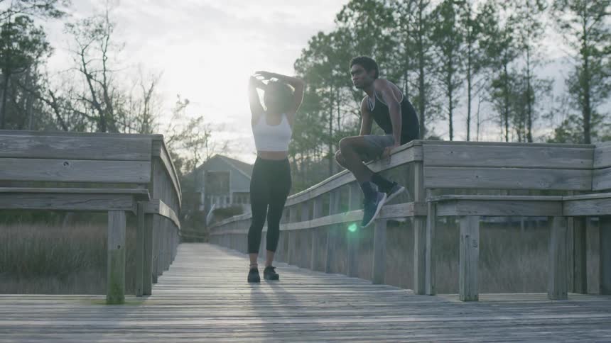 Back lit by the sun a woman stretches on a wooden boardwalk while a man sits on the railing beside her
