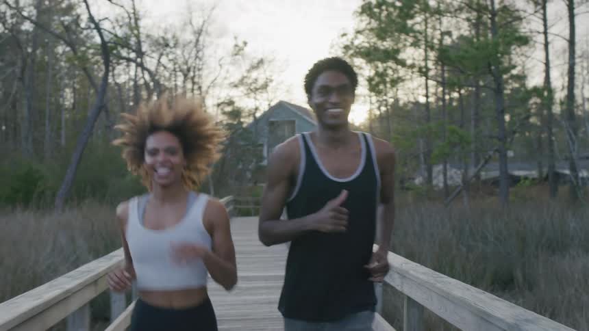 Sun shines past a house on a cloudy day onto two people running across a wooden footbridge