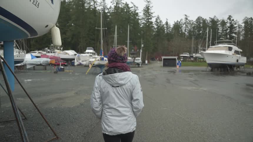 Following from behind a person wearing a winter hat and jacket walks through a boatyard filled with boats stored on land