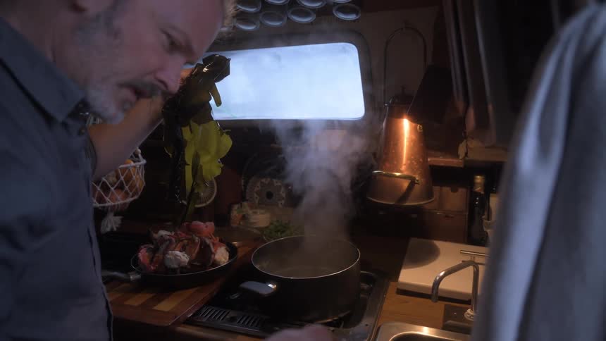 A man cooks in a dimly lit boat galley adding ingredients to a steaming pot next to a pan of cooked crab