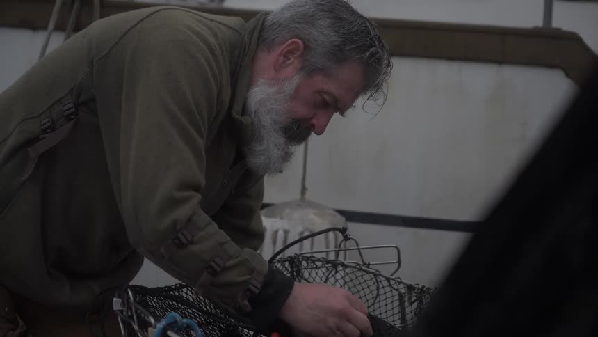 Older man works on the mesh of a trap while on a dock next to a boat