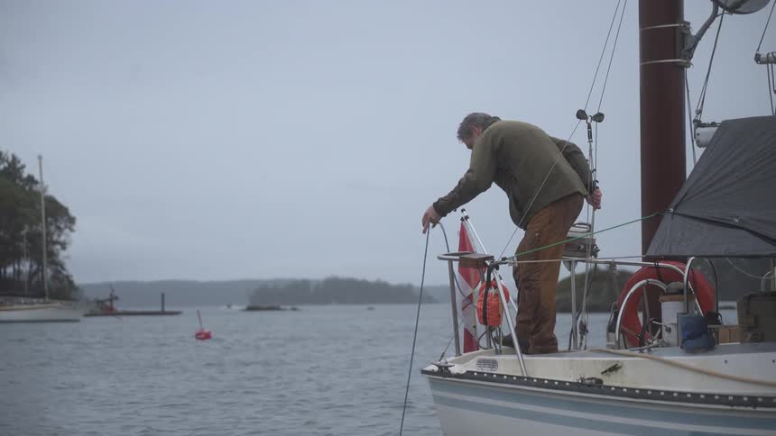 A fisherman on a boat lowers a crab trap into the water with buoys and a distant shoreline visible in the background