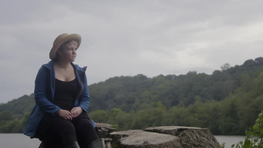 A young woman is sitting on a tree trunk gazing out at a river on a cloudy day