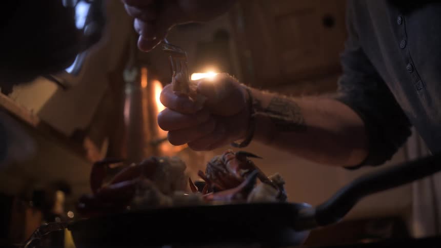Using a fork hands carefully extract cooked meat from a crab claw resting in a pan in a lowlight kitchen