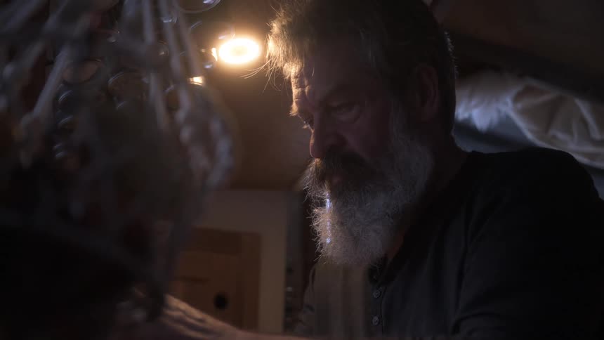 A focused bearded man works intently under a single overhead light in a dimly lit room
