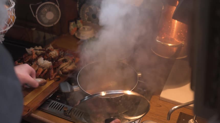 A bearded person uses a phone to time as they place crab legs into a steaming pot on a stovetop