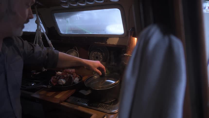 A man cooks crab in a steaming pot on a stovetop in a cramped boat kitchen