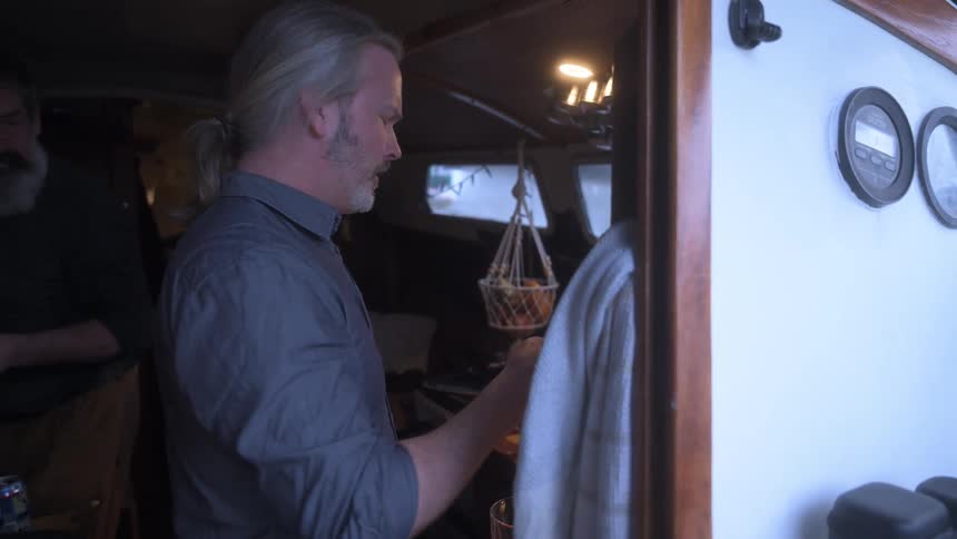 Two people in a dimly lit boat cabin engage in pouring a drink from a bottle of wine into a glass