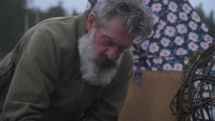 A closeup of a person kneeling beside a round handcrafted basket made of fresh twigs with an umbrella resting nearby in a tranquil natural setting