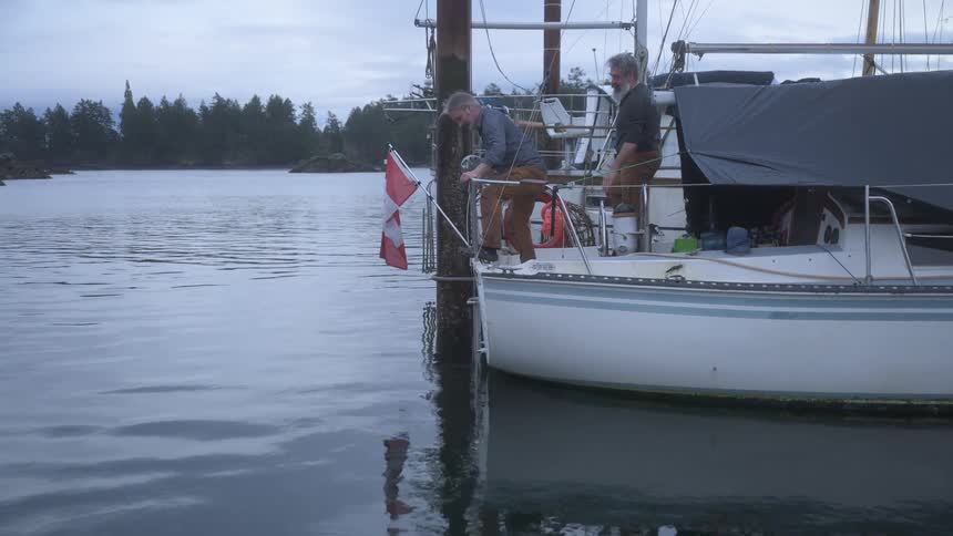Two fishermen stand on a sailboat docked at the pier pulling a rope from the lake as a canadian flag flutters nearby