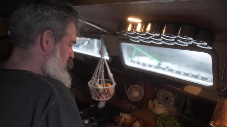 A person stands in a small kitchen beside hanging baskets of fruit and spice jars with a window view of a boat deck