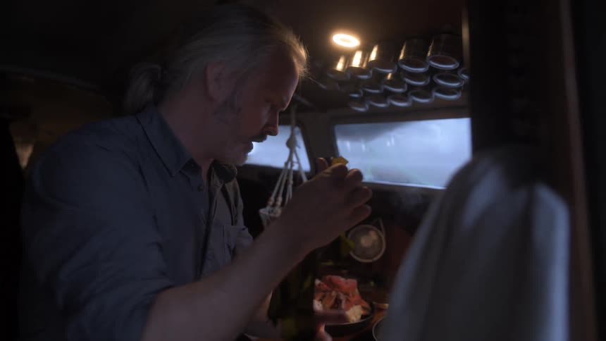 A man prepares crab legs in a cozy boat kitchen breaking fresh kelp leaves with his hands amidst a warm and rustic atmosphere