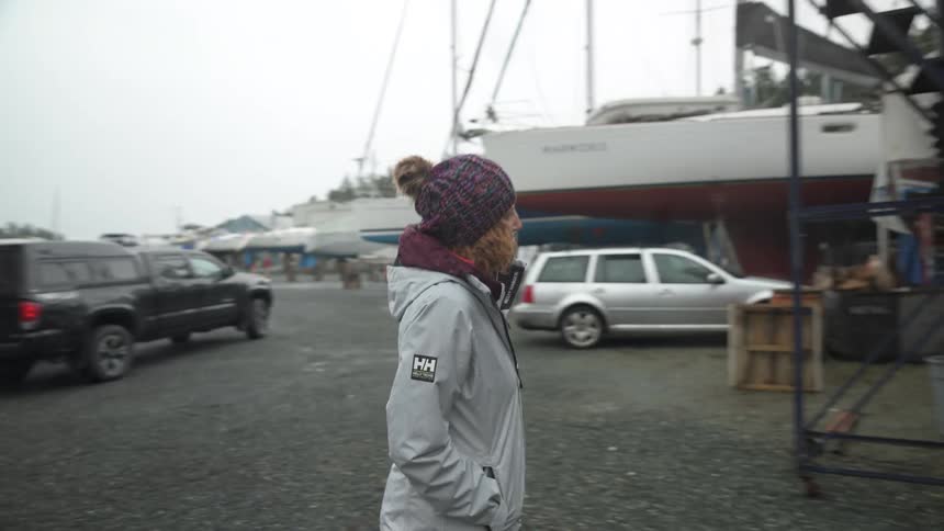 A woman walks toward several boats raised on land with trees in the background wearing a white jacket and a vibrant knit hat