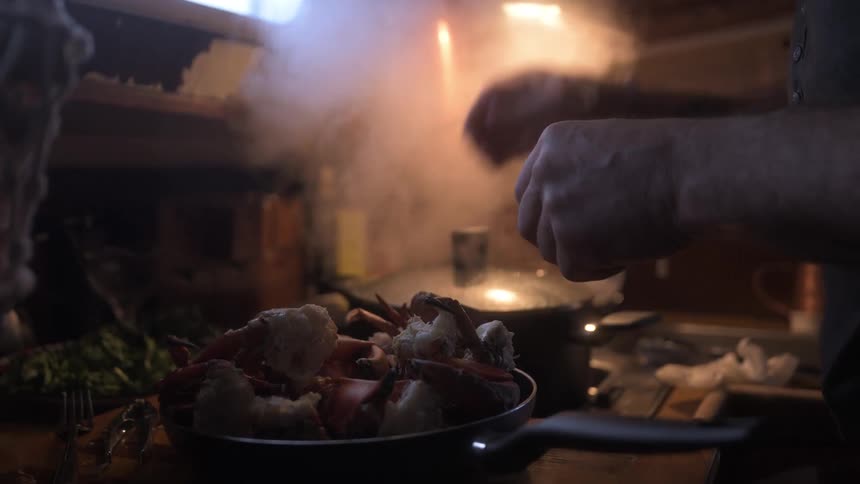 A person lifts crab legs from a pan on the stovetop in a dimly lit kitchen and transfers them into a steaming pot