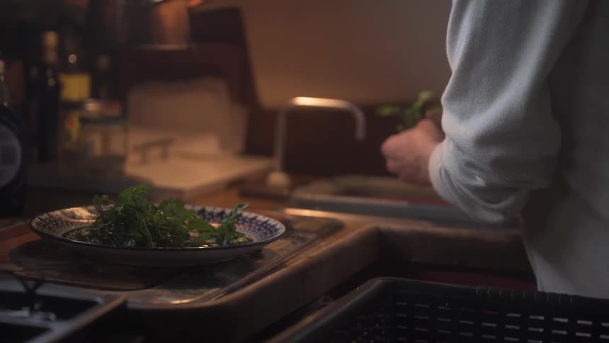 A person carefully cleans fresh herbs and places them on a decorative plate in a dimly lit kitchen