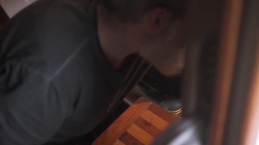 A man uses a match to ignite a stovetop flame and places a pot filled with water in a cramped kitchen observed through a window from outside
