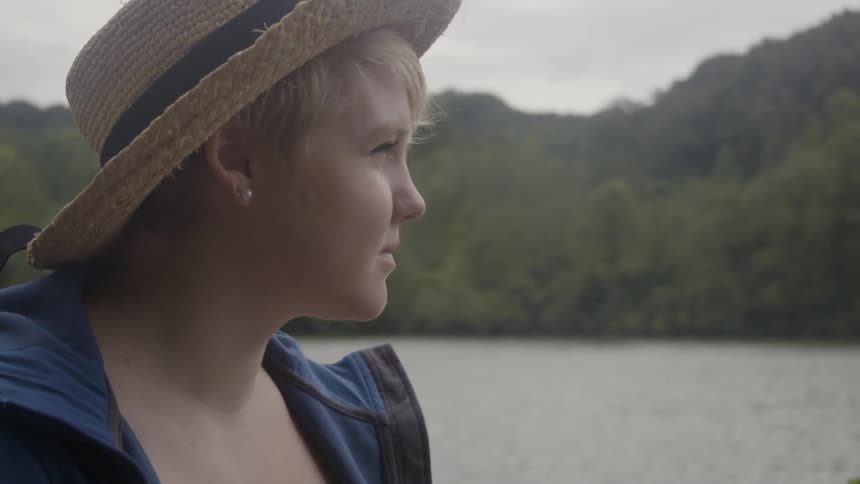 Blonde woman wearing a hat looking forward towards the large river beside her