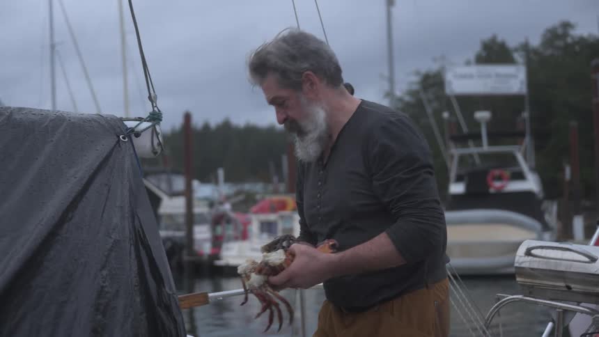 A bearded man on a boat holds crabs while other boats and a dock are visible in the cloudy background
