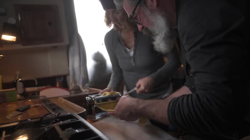 Two individuals operate a pasta machine rolling fresh dough in a home kitchen