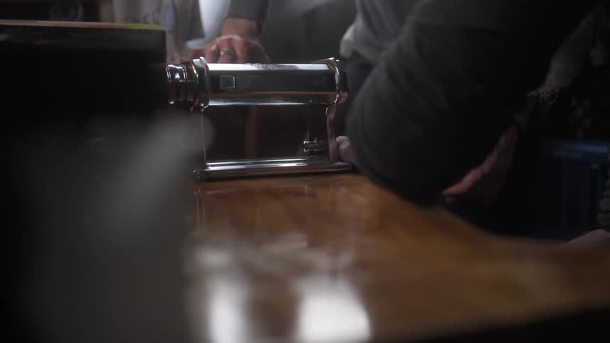 A shiny metal pasta machine sits on a wooden table as two people work together to adjust and secure it in place
