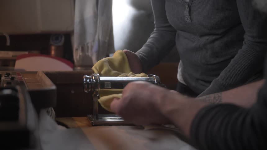 A couple operate a pasta machine rolling fresh dough in a home kitchen