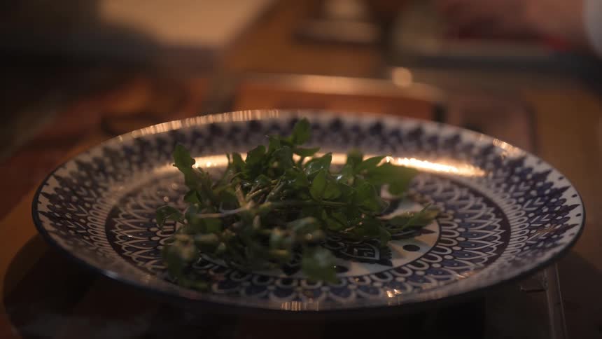 A person places small bunch of fresh green herbs likely watercress on a blue and white patterned plate over a wooden table