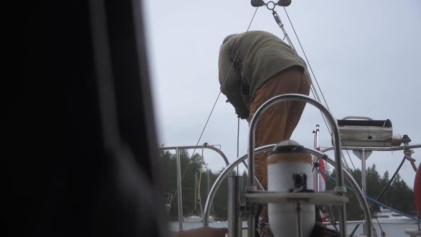 The back of a man is visible as he lowers something with a rope from the side of a boat viewed from inside the vessel on an overcast day