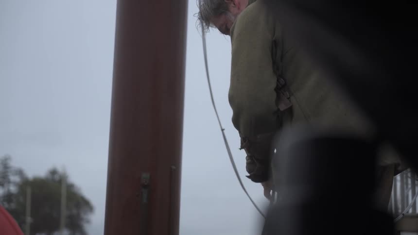 A man on a boat unties and handles a rope next to the mast on a cloudy day