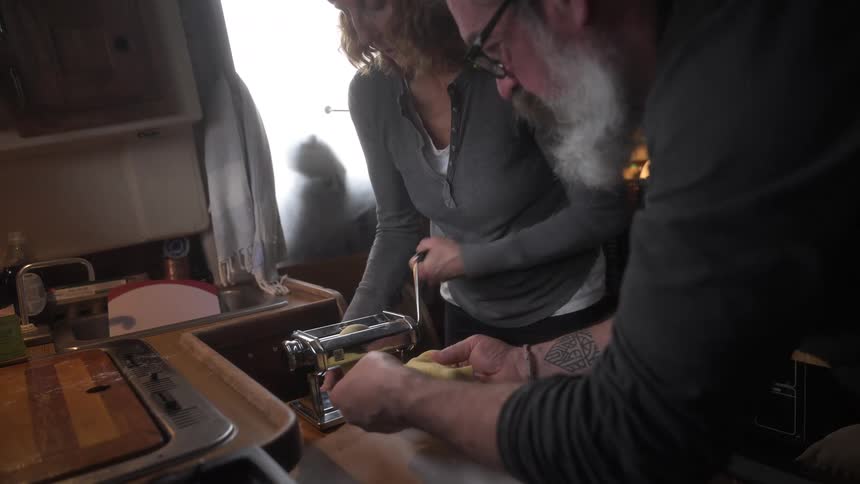A man and woman work together using a pasta maker in a small kitchen space