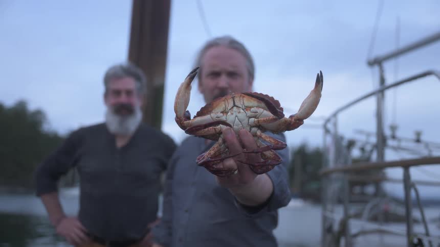 Two men are standing near boats as one shows off a big crab